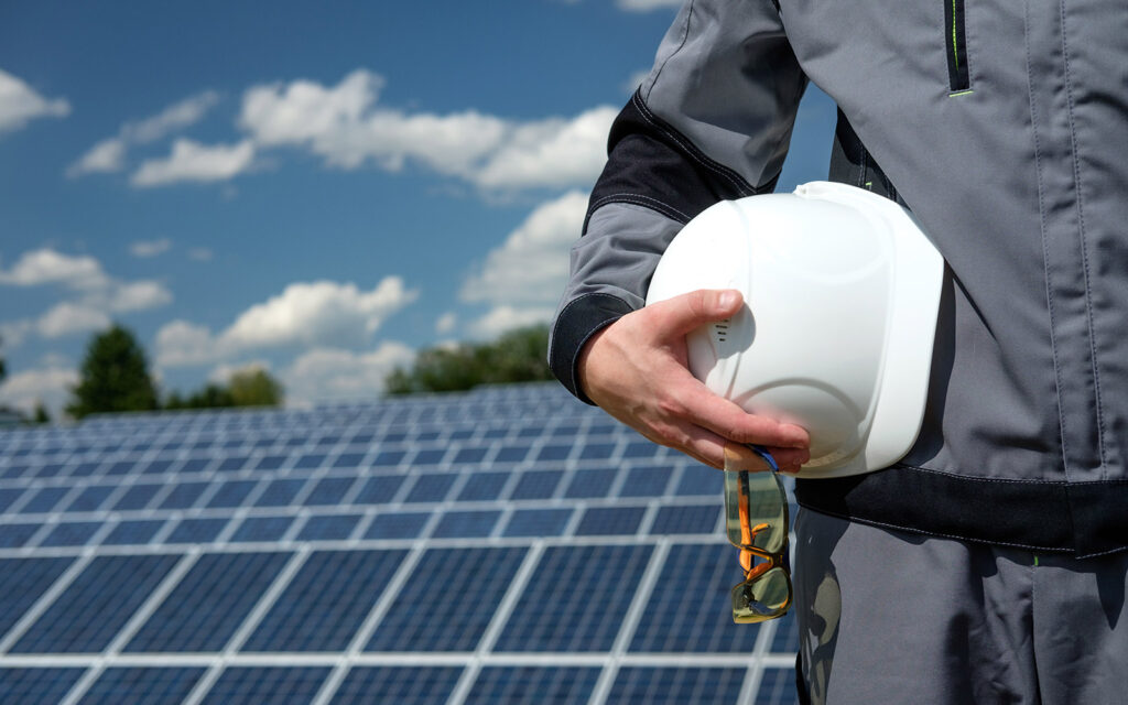 A contractor holding a hardhat and safety glasses in front of a field of solar panels.