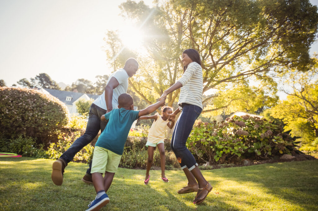 A family holding hands and dancing in their backyard with the sun shining through the trees.