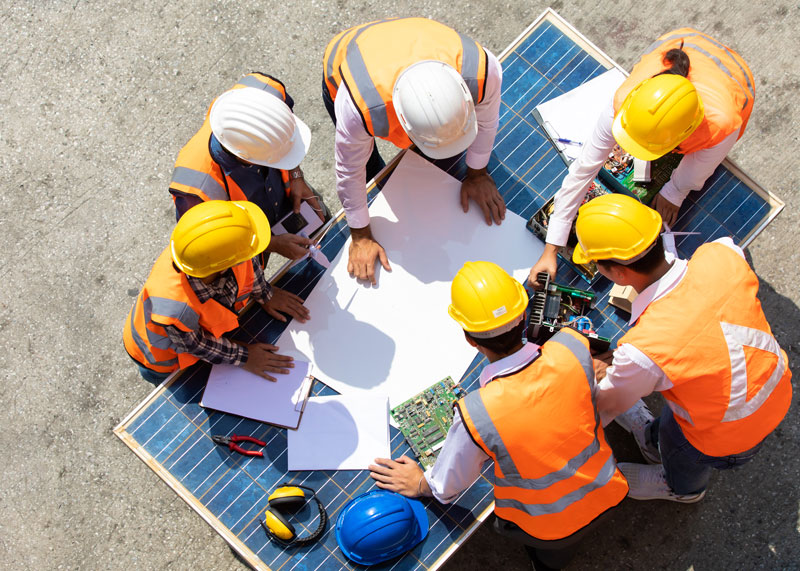 A Group of people wearing hi-visibility vests and hard hats huddled around papers and tools on a solar panel.